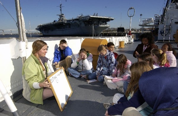 Marine science instructor conducts class on USCG Ingham with USS Yorktown (CV-10) in background.