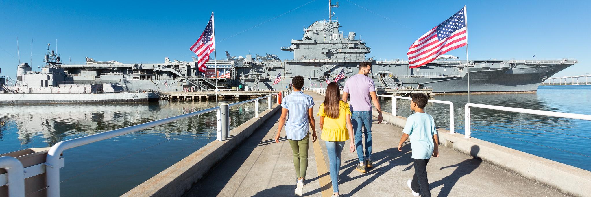 A family walks down the pathway toward the USS Yorktown aircraft carrier at Patriots Point, surrounded by American flags and calm waters. 