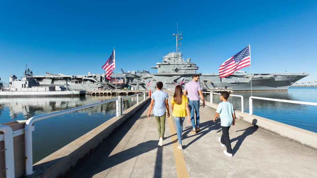 A family walks down the pathway toward the USS Yorktown aircraft carrier at Patriots Point, surrounded by American flags and calm waters. 