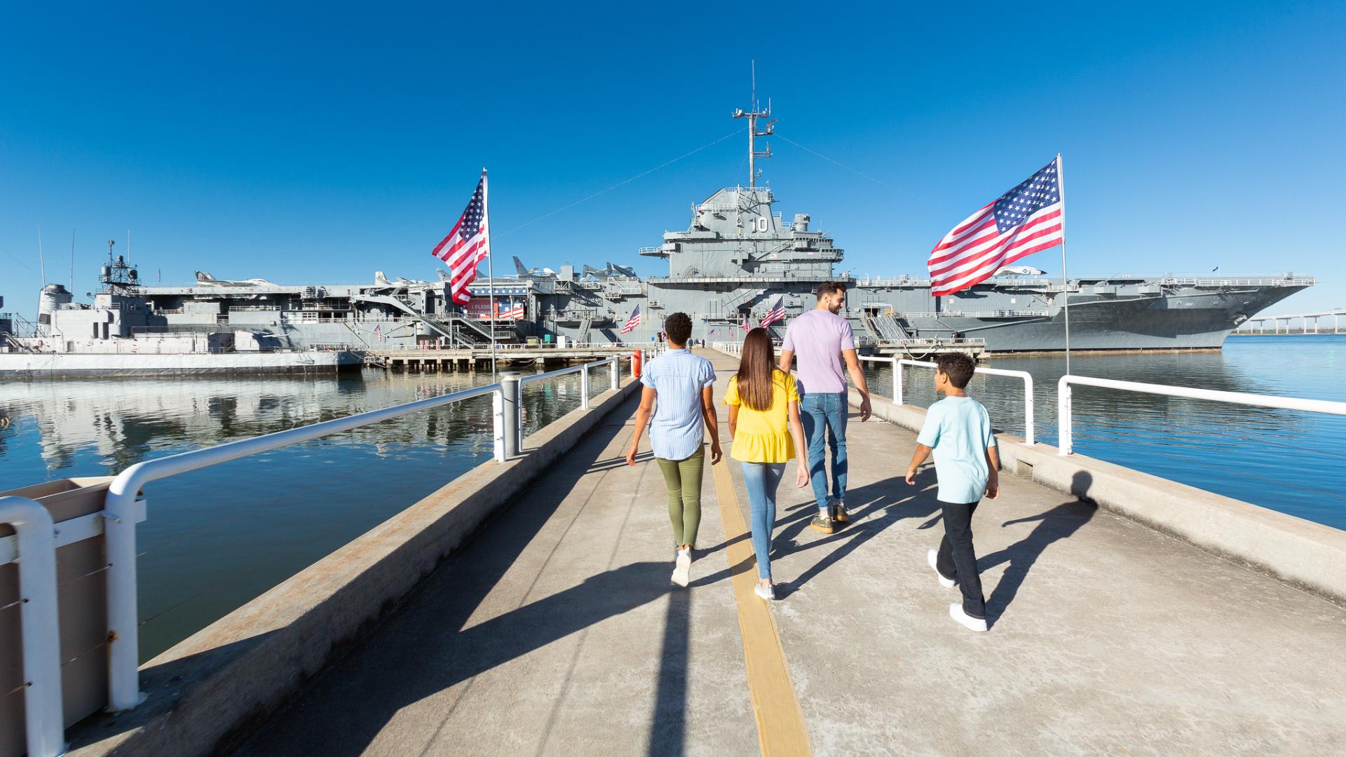 A family walks down the pathway toward the USS Yorktown aircraft carrier at Patriots Point, surrounded by American flags and calm waters. 