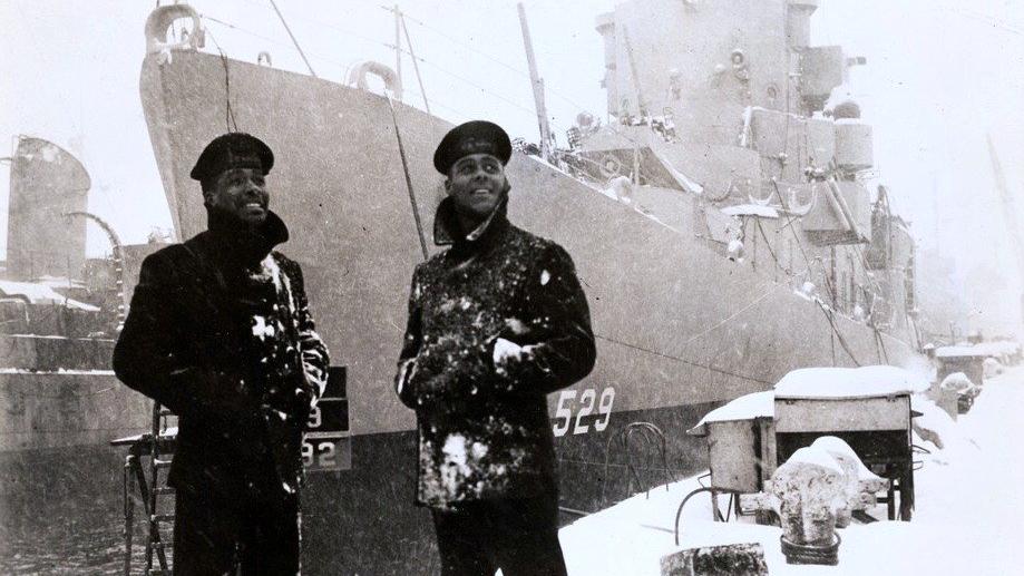 Two black sailors dressed in winter jackets stand in front of the USS Mason, with the numbers 529 on the ship, in the snow.