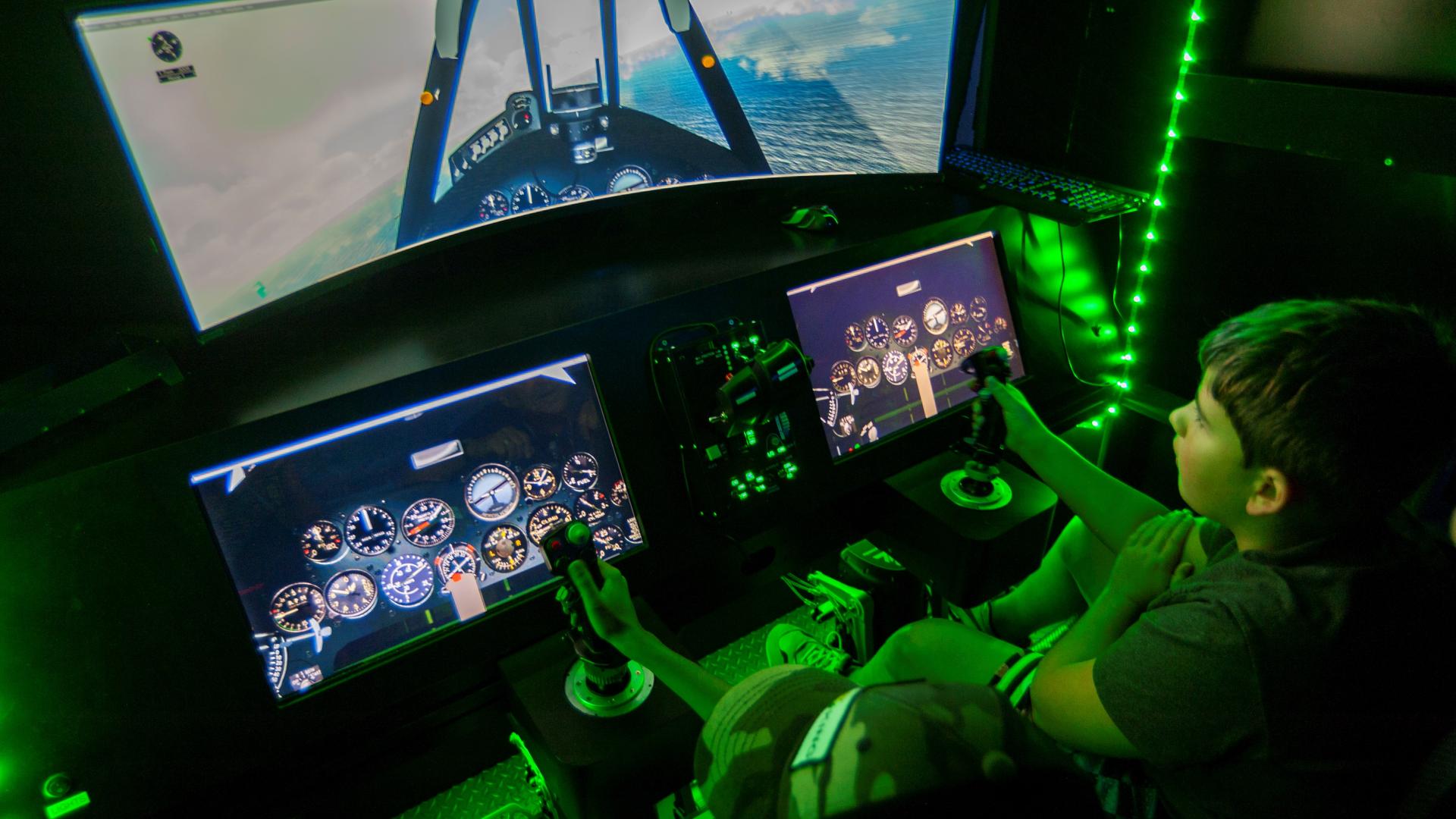 Child uses handheld joystick to virtually fly military aircraft illuminated by green lights.