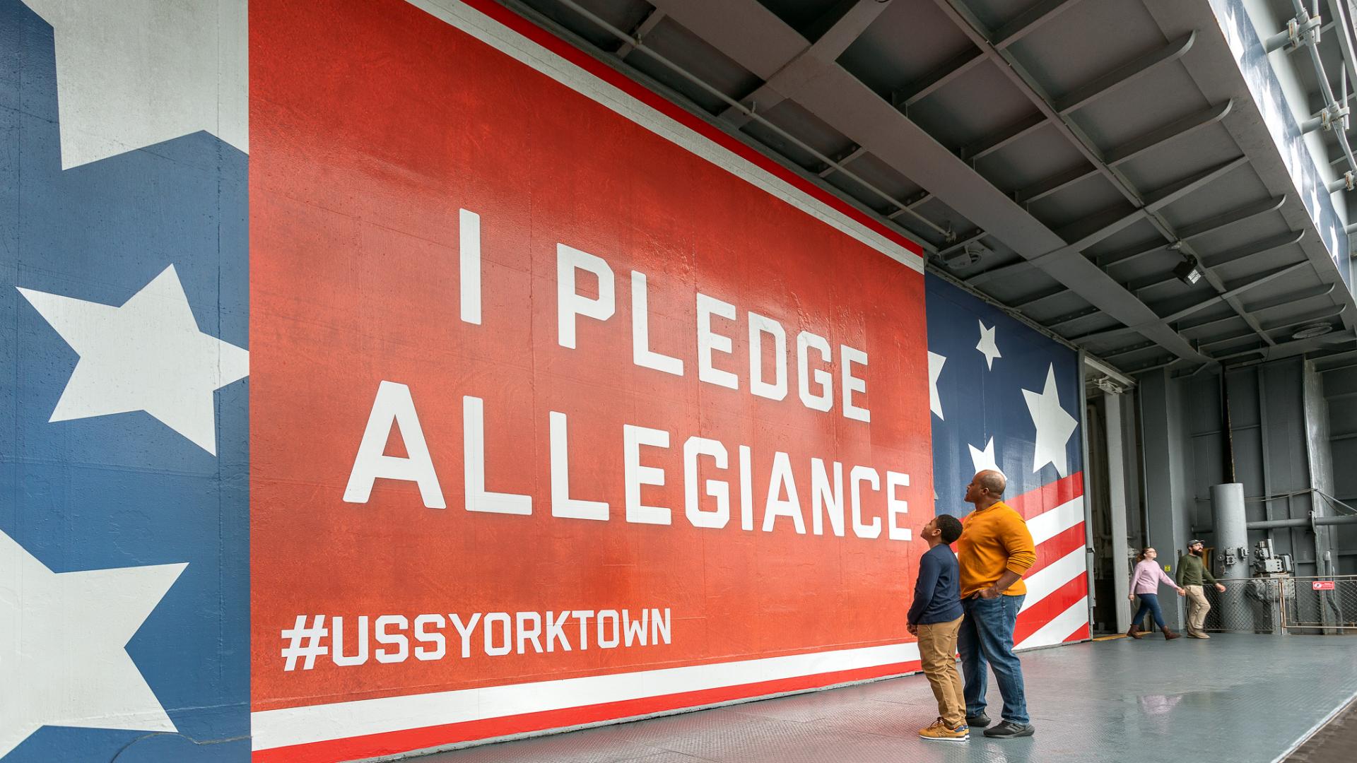 Father and son stand in front of I Pledge Allegiance sign on hangar bay door. 
