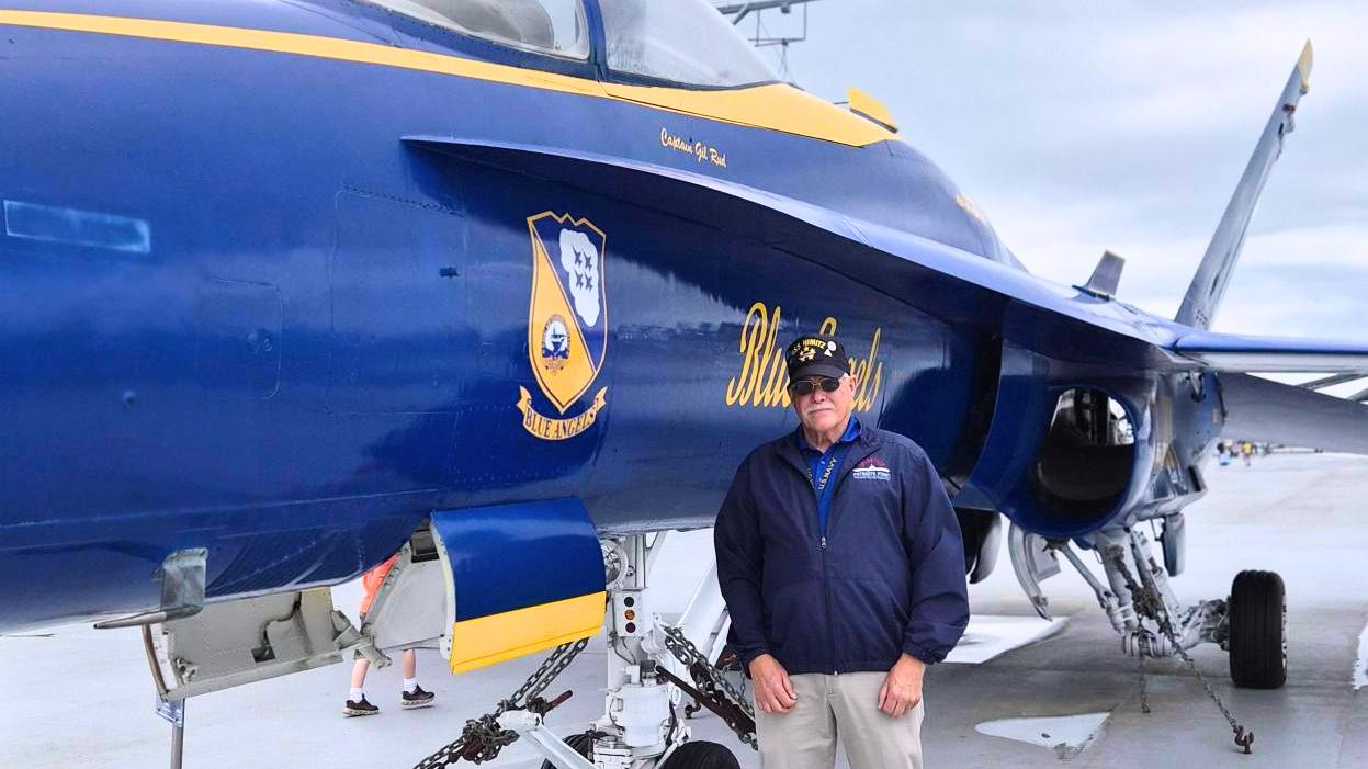 Painted aircraft with 'Blue Angel' on aircraft carrier flight deck and veteran Tom Simons in front of it.
