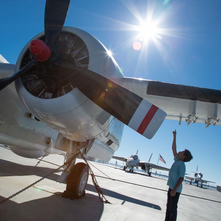 a small boy stands next to a large aircraft pointing to the wing above his head