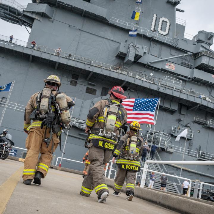 Firefighters in full gear walk toward the USS Yorktown, with an American flag prominently displayed.