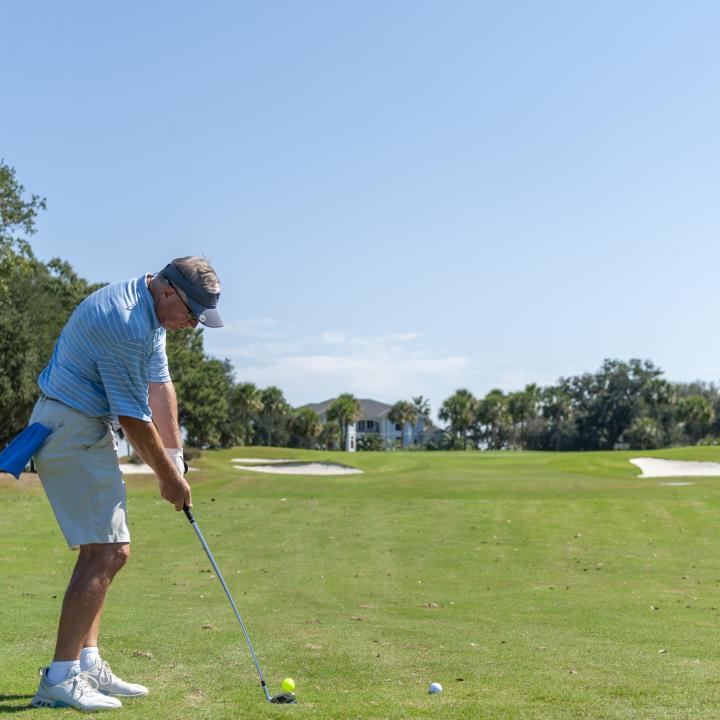A golfer prepares to take a swing on a sunny day, standing on a well-maintained golf course surrounded by trees and distant homes.