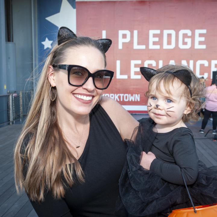 A mom and daughter dress up as cats for Halloween while standing in front of a red, white, and blue banner on the USS Yorktown (CV-10) that says "I pledge allegiance." 