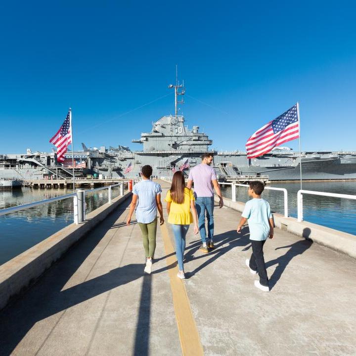 A family walks down the pathway toward the USS Yorktown aircraft carrier at Patriots Point, surrounded by American flags and calm waters. 