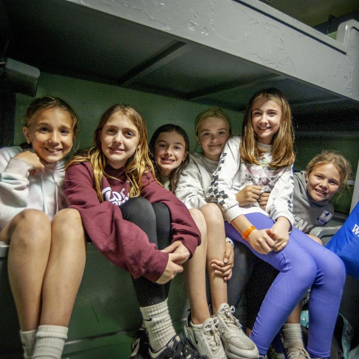 Six girls sit on a bunk together smiling.