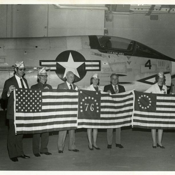 This photograph shows a group of men and women posing with flags for the United States Bicentennial on deck for opening day ceremonies aboard the USS Yorktown, January 3, 1976.