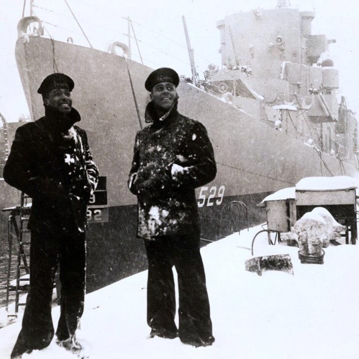 Two black sailors dressed in winter jackets stand in front of the USS Mason, with the numbers 529 on the ship, in the snow.