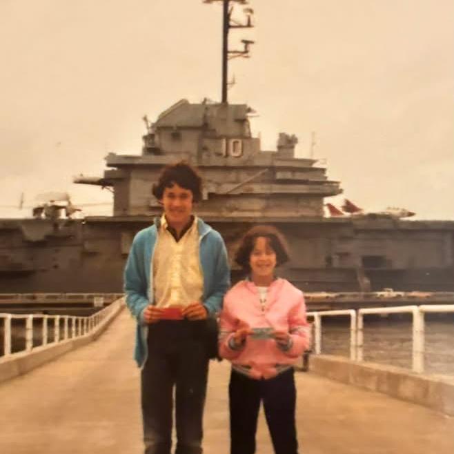 A boy in blue jacket and girl in pink shirt stand in front of the ship USS Yorktown with the number 10 on the side.