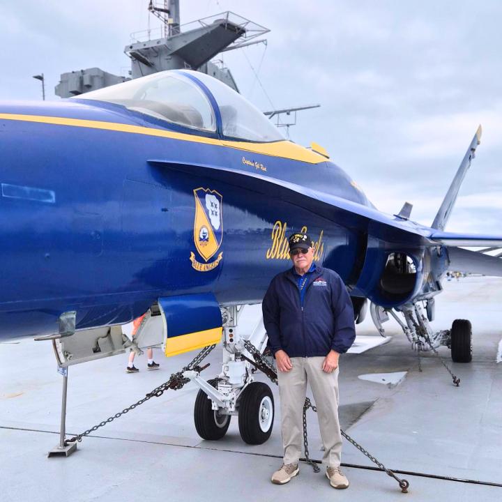 Painted aircraft with 'Blue Angel' on aircraft carrier flight deck and veteran Tom Simons in front of it.