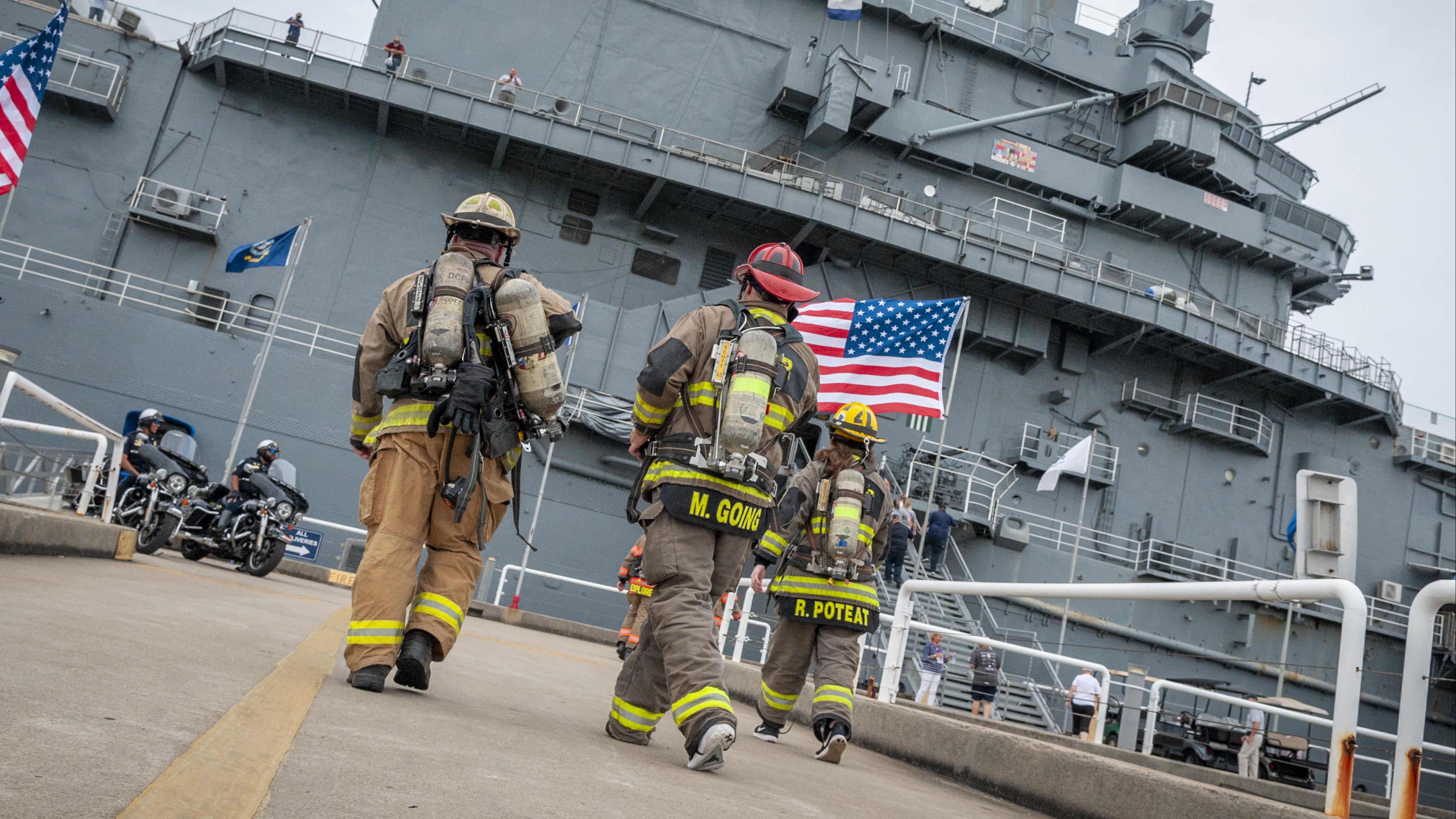 Firefighters in full gear walk toward the USS Yorktown, with an American flag prominently displayed.