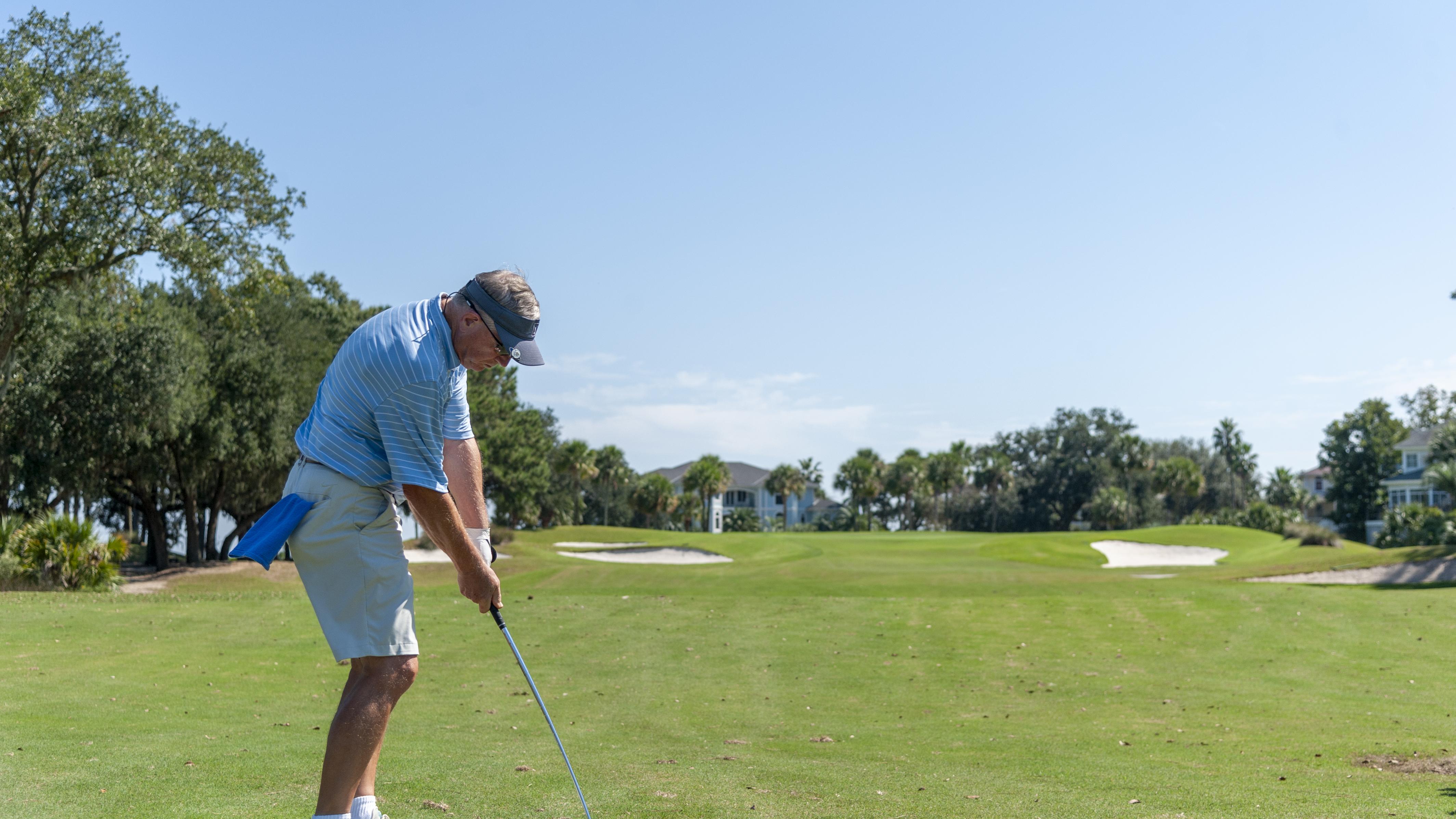 A golfer prepares to take a swing on a sunny day, standing on a well-maintained golf course surrounded by trees and distant homes.