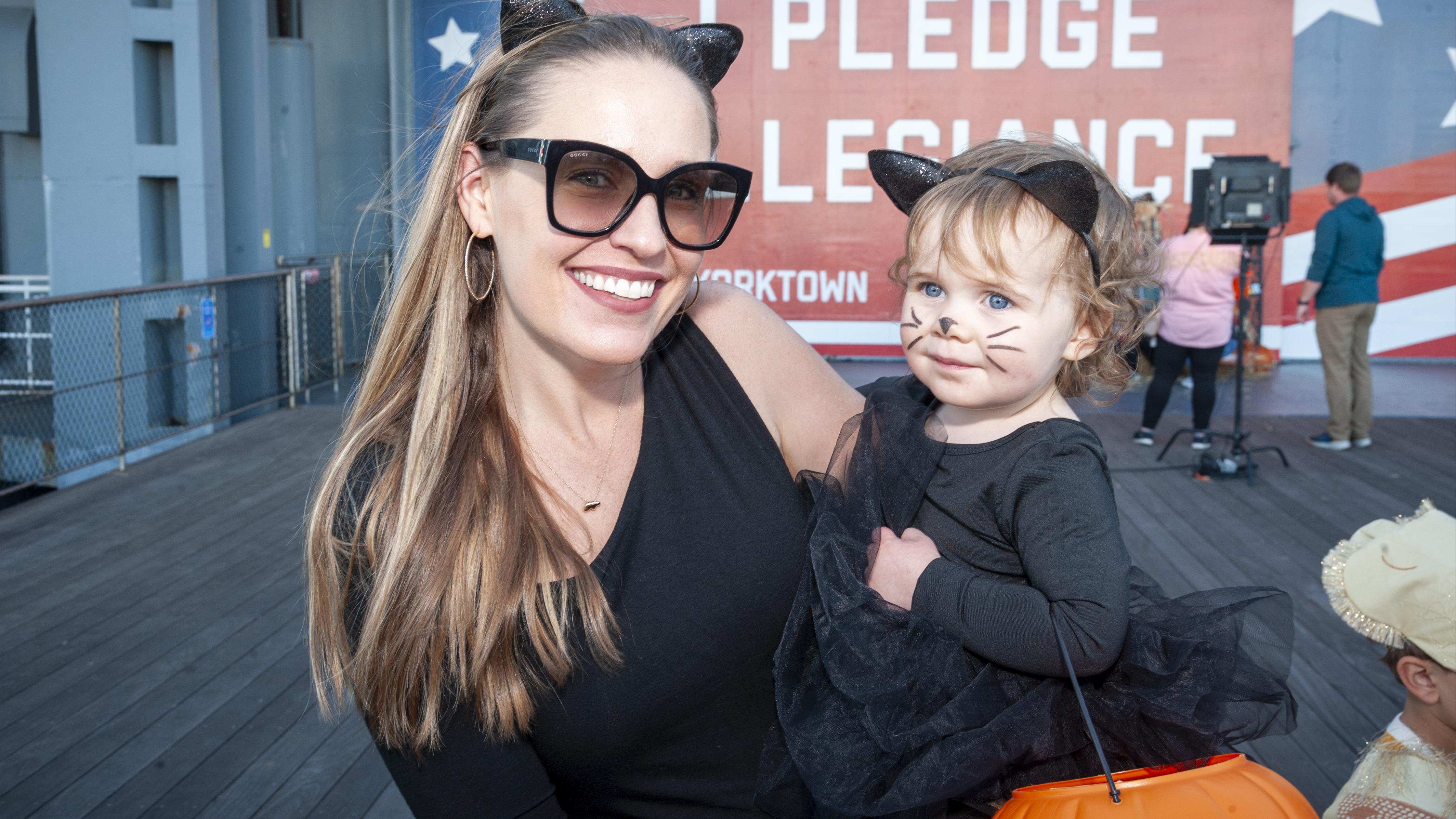 A mom and daughter dress up as cats for Halloween while standing in front of a red, white, and blue banner on the USS Yorktown (CV-10) that says "I pledge allegiance." 