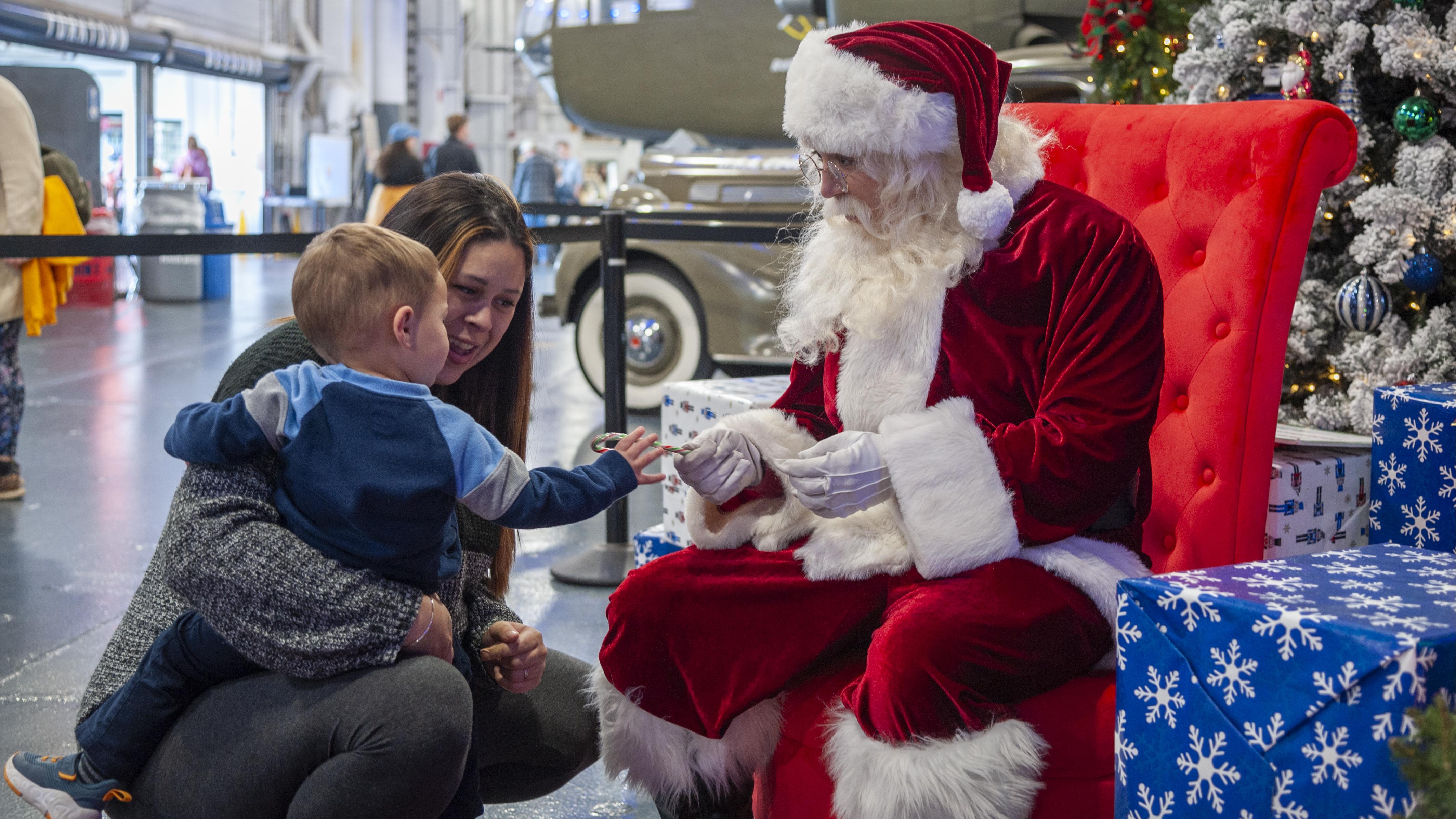 A young child and their parent interact with Santa Claus, who is seated in a red chair and handing the child a candy cane, surrounded by festive holiday decorations and gifts.