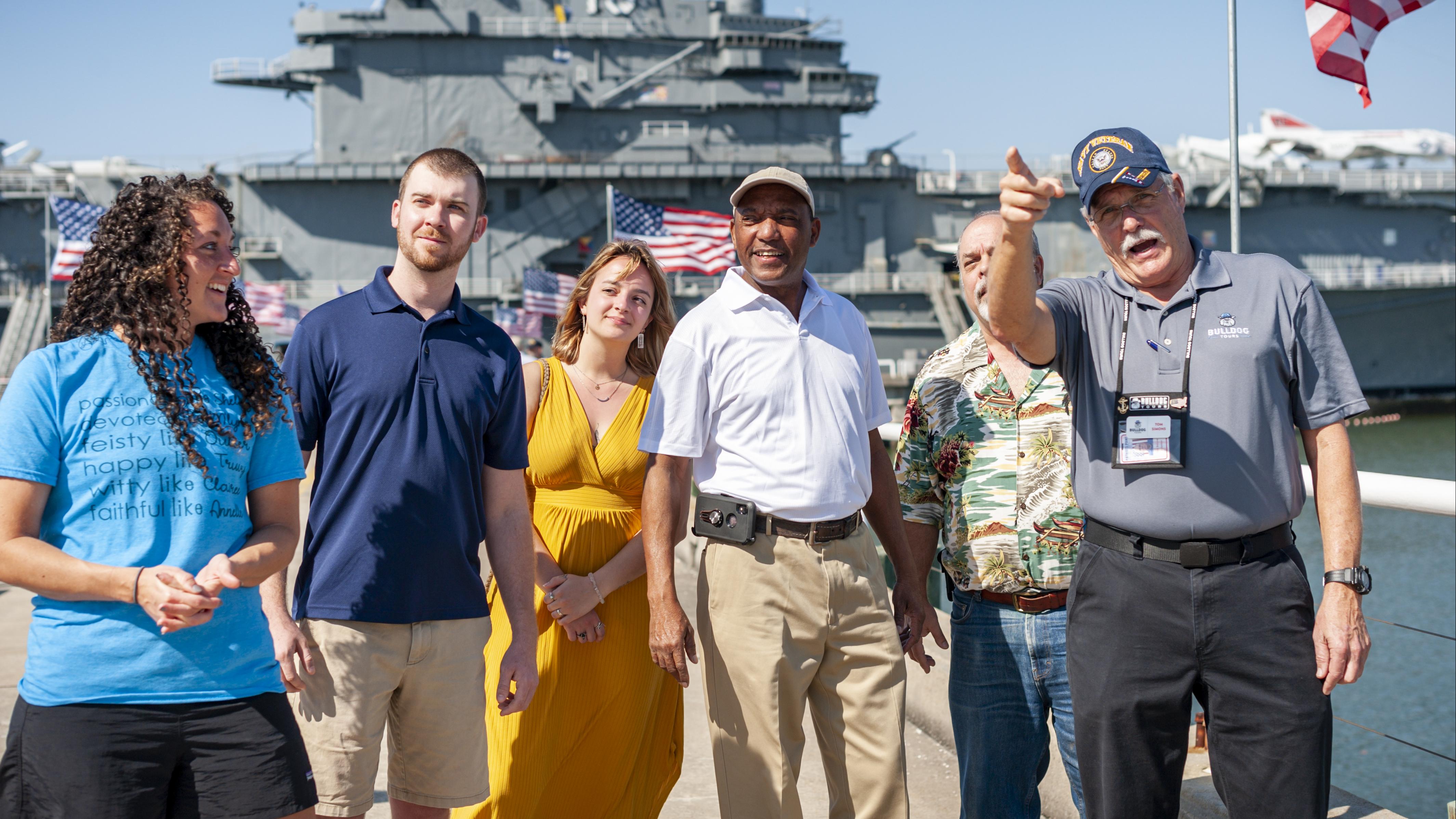 A group of visitors listen to a tour guide while on the pier near the USS Yorktown. 