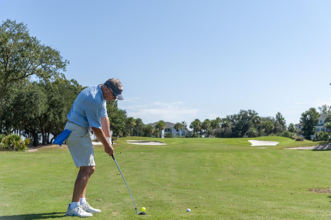 A golfer prepares to take a swing on a sunny day, standing on a well-maintained golf course surrounded by trees and distant homes.