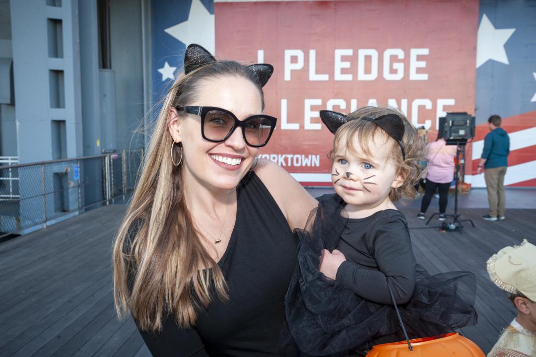 A mom and daughter dress up as cats for Halloween while standing in front of a red, white, and blue banner on the USS Yorktown (CV-10) that says "I pledge allegiance." 