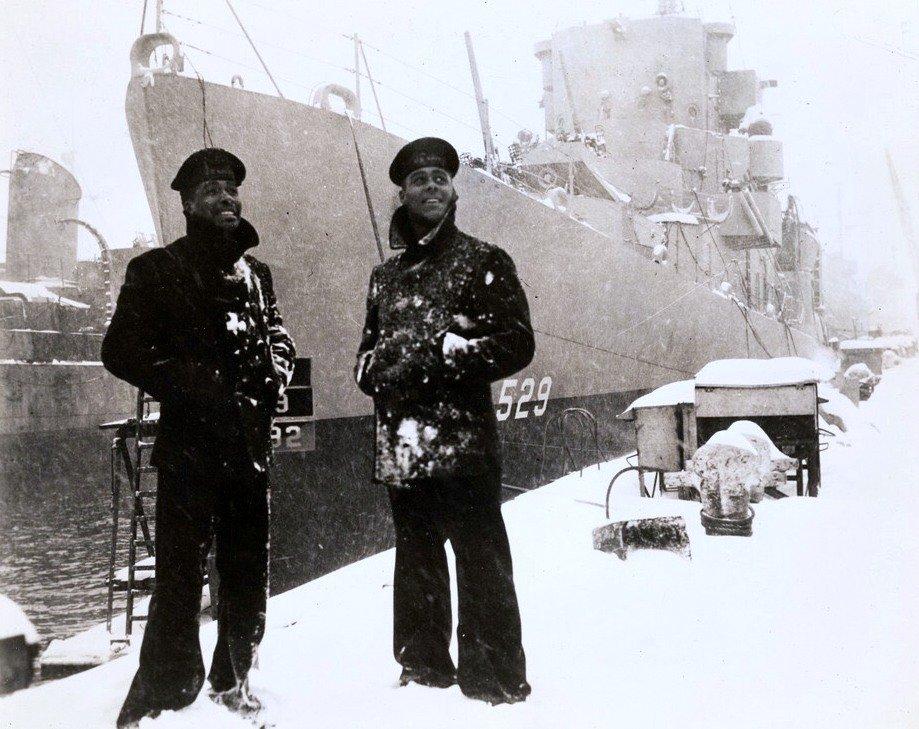 Two black sailors dressed in winter jackets stand in front of the USS Mason, with the numbers 529 on the ship, in the snow.