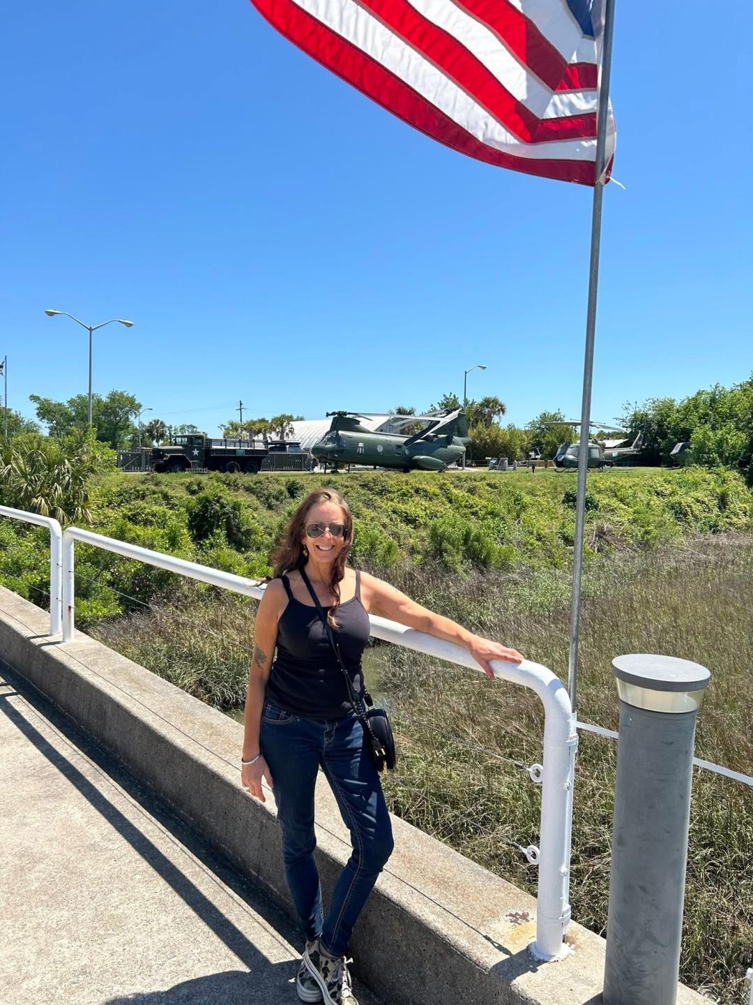 Woman stands in black top and blue jeans next to an American flag with a sunny background.